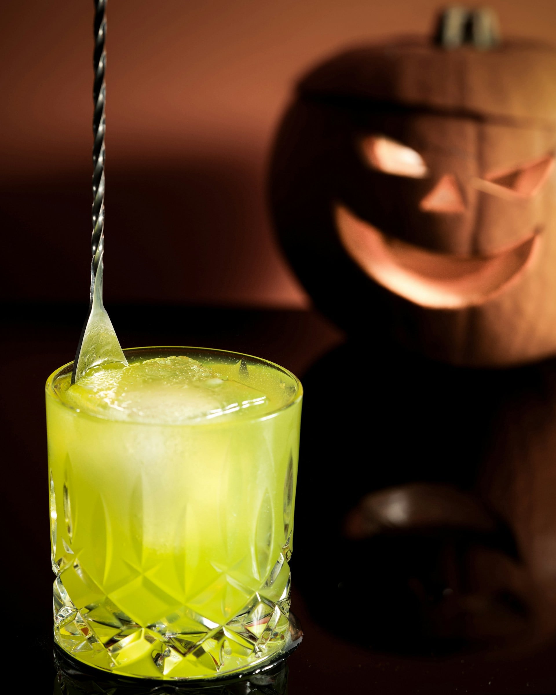 Bright green cocktail in a cut-crystal glass with a lit carved pumpkin glowing in the background