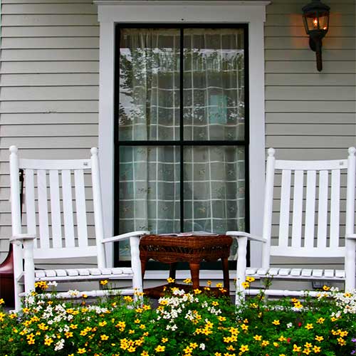 2 white rocking chairs on a front porch with yellow flowers and an accent table
