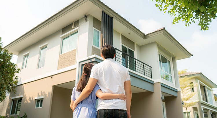 Back portrait of young couple standing and hugging together looking happy in front of their new house to start new life. Family, age, home, real estate and people concept.