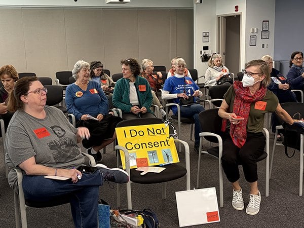 DeFlock Fairfax advocates seated at a Fairfax County meeting, one holding an 'I Do Not Consent' sign