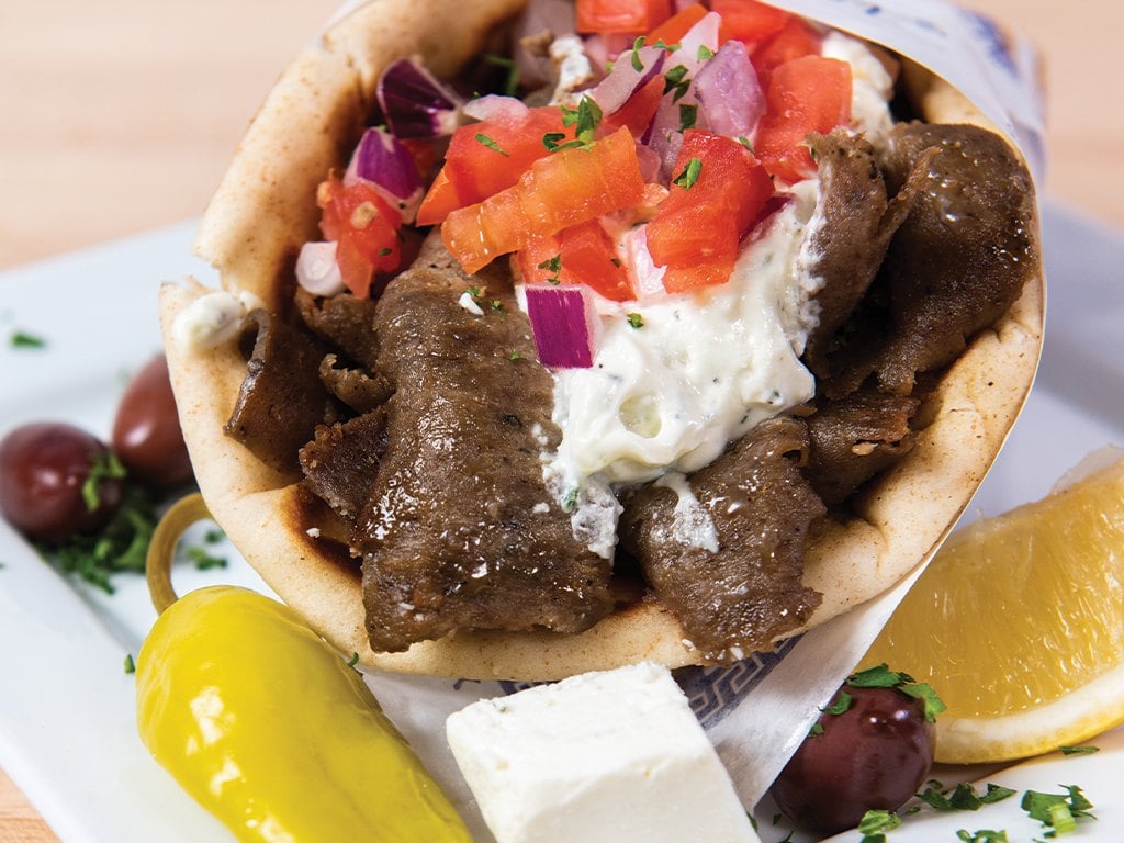 A gyro pita with shaved meat, tzatziki, tomato and red onion, served with feta, olives and a lemon wedge