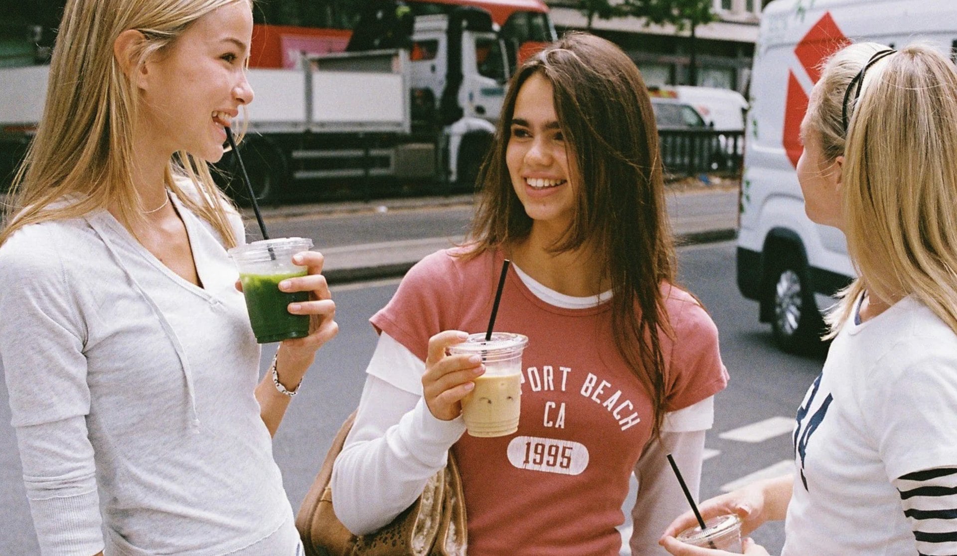 Three young women in Brandy Melville tops holding iced drinks on a city sidewalk