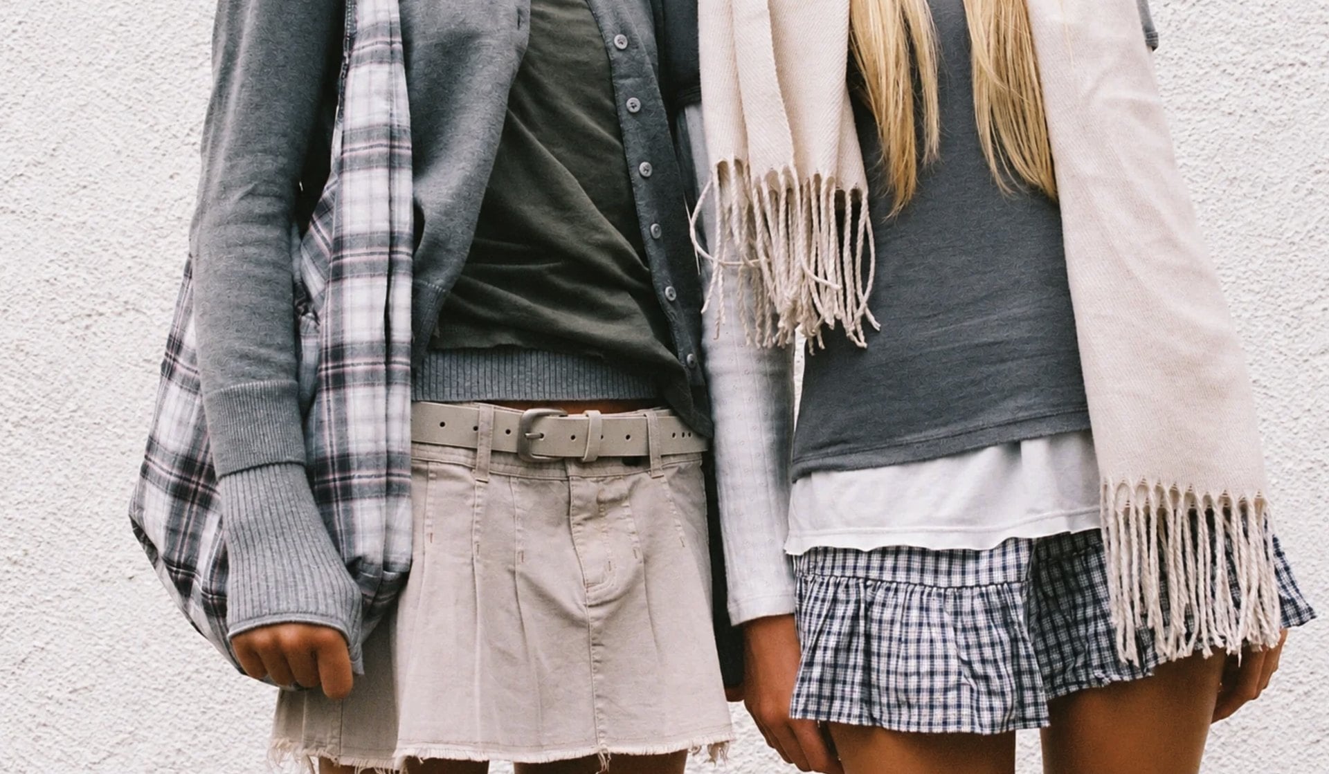 Two people in Brandy Melville mini skirts, cardigans and a fringed scarf against a white wall