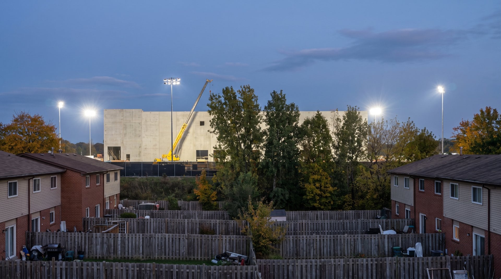 Illustration of townhouse backyards in Reston with a strip of trees and a large data center under construction behind them at dusk