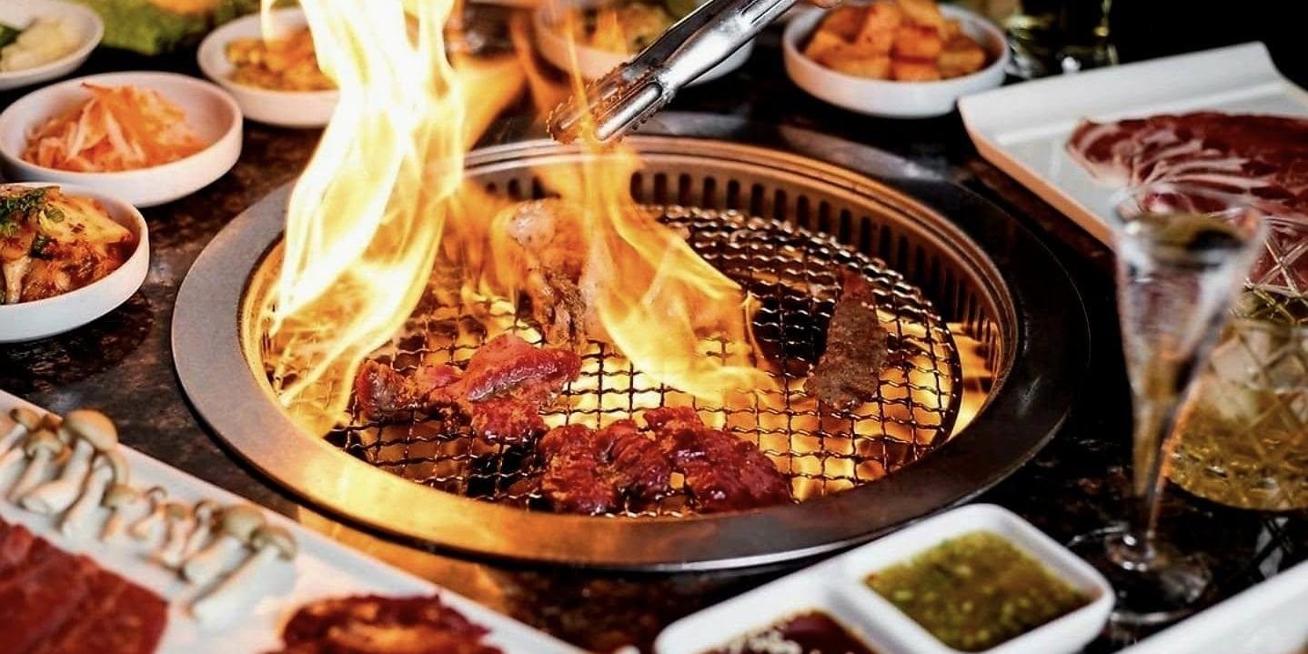 Marinated meat cooking over flames on a tabletop Korean barbecue grill surrounded by banchan side dishes at H Pot in Herndon