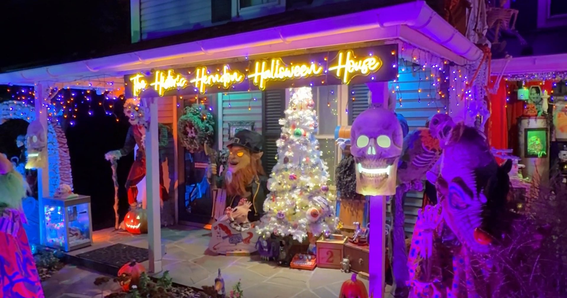 The porch of the Historic Herndon Halloween House on Spring Street at night, lit purple, with a neon sign, a white Christmas tree and a glowing skull