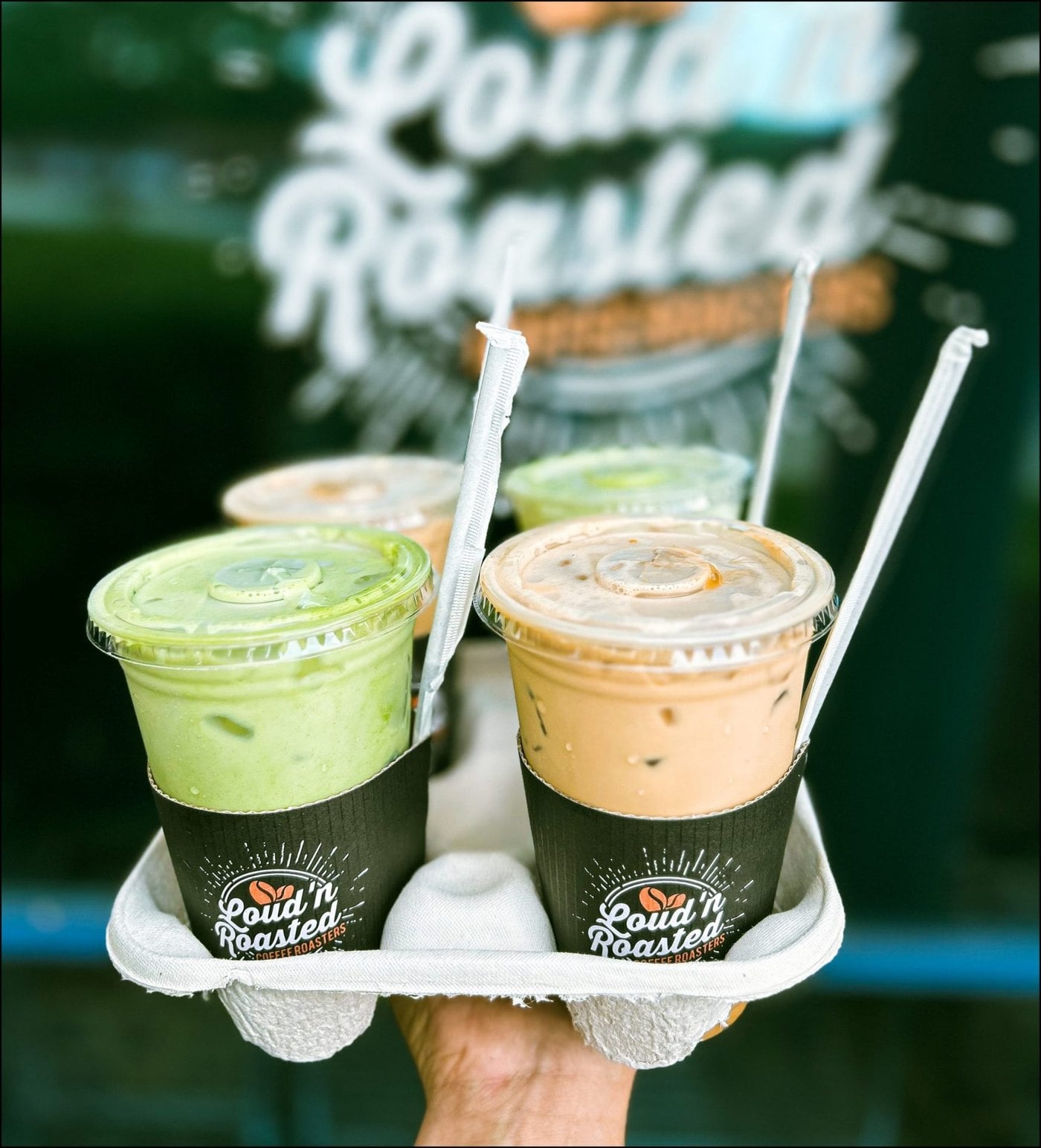 A hand holding a carrier of iced matcha and iced lattes in Loud'n Roasted sleeves in front of the shop's logo window