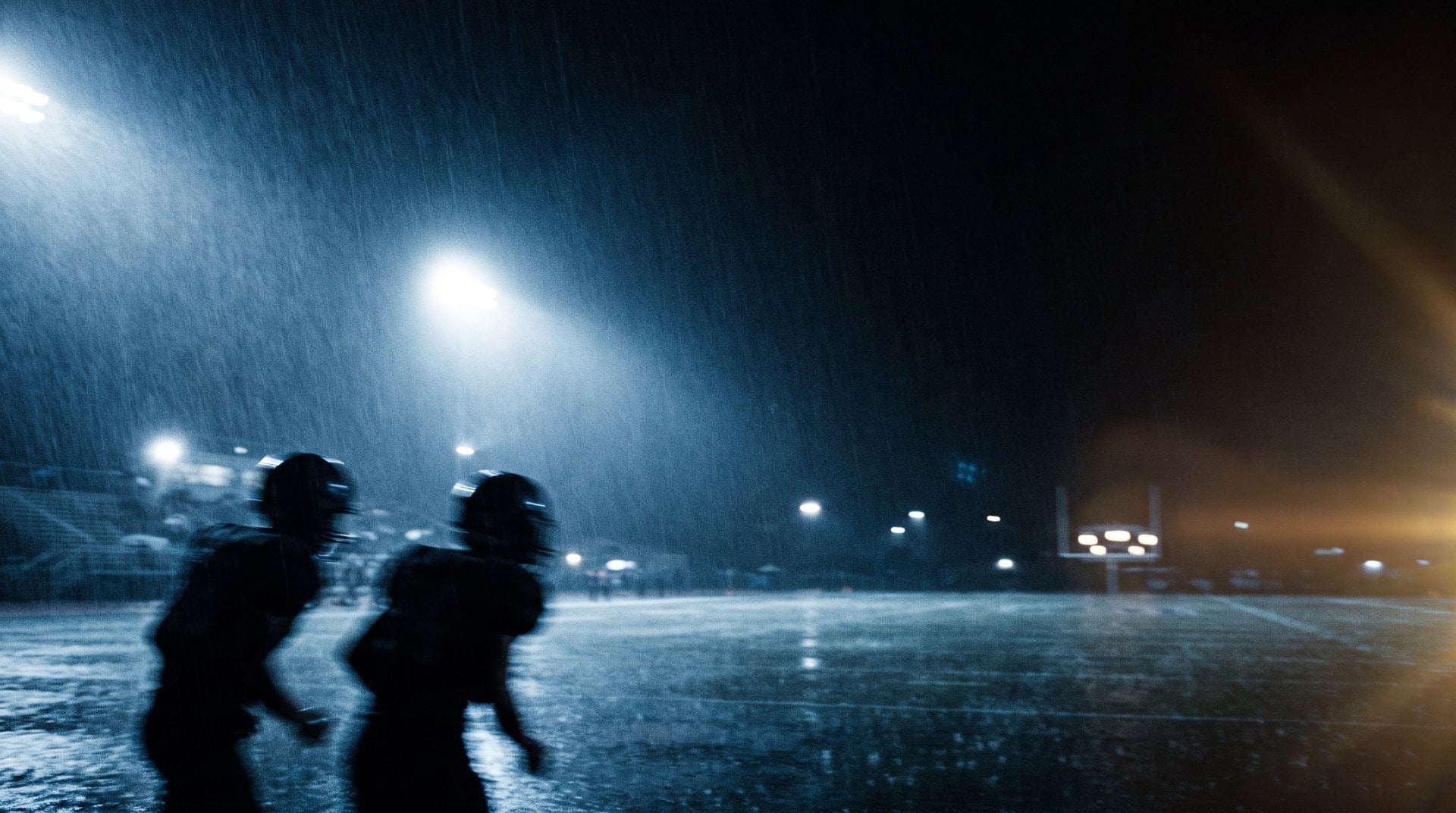 Blurred silhouettes of two football players running through rain under stadium lights