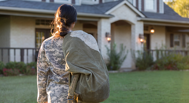 Young woman in military uniform faces her back towards the camera, as she takes one last look at her home before leaving. She has a backpack over her shoulder.
