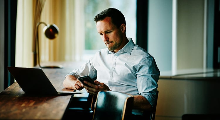 Businessman looking at smartphone while sitting at counter in office coworking space working on project on laptop