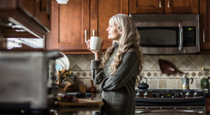 Older woman in her kitchen looking out the window with a cup of tea