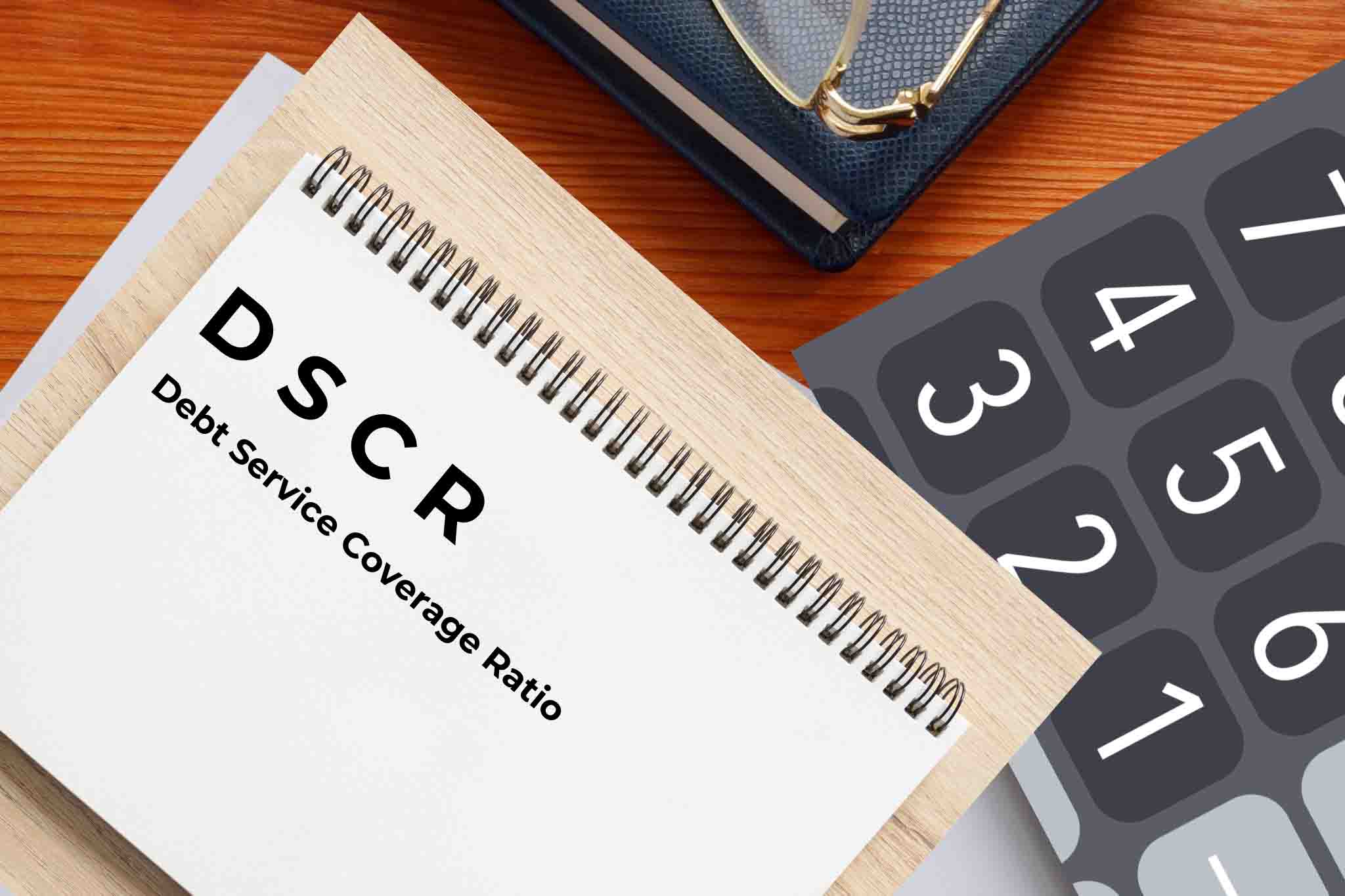 A notepad with the text 'DSCR Debt Service Coverage Ratio' on a wooden desk, next to a calculator and a pair of glasses.