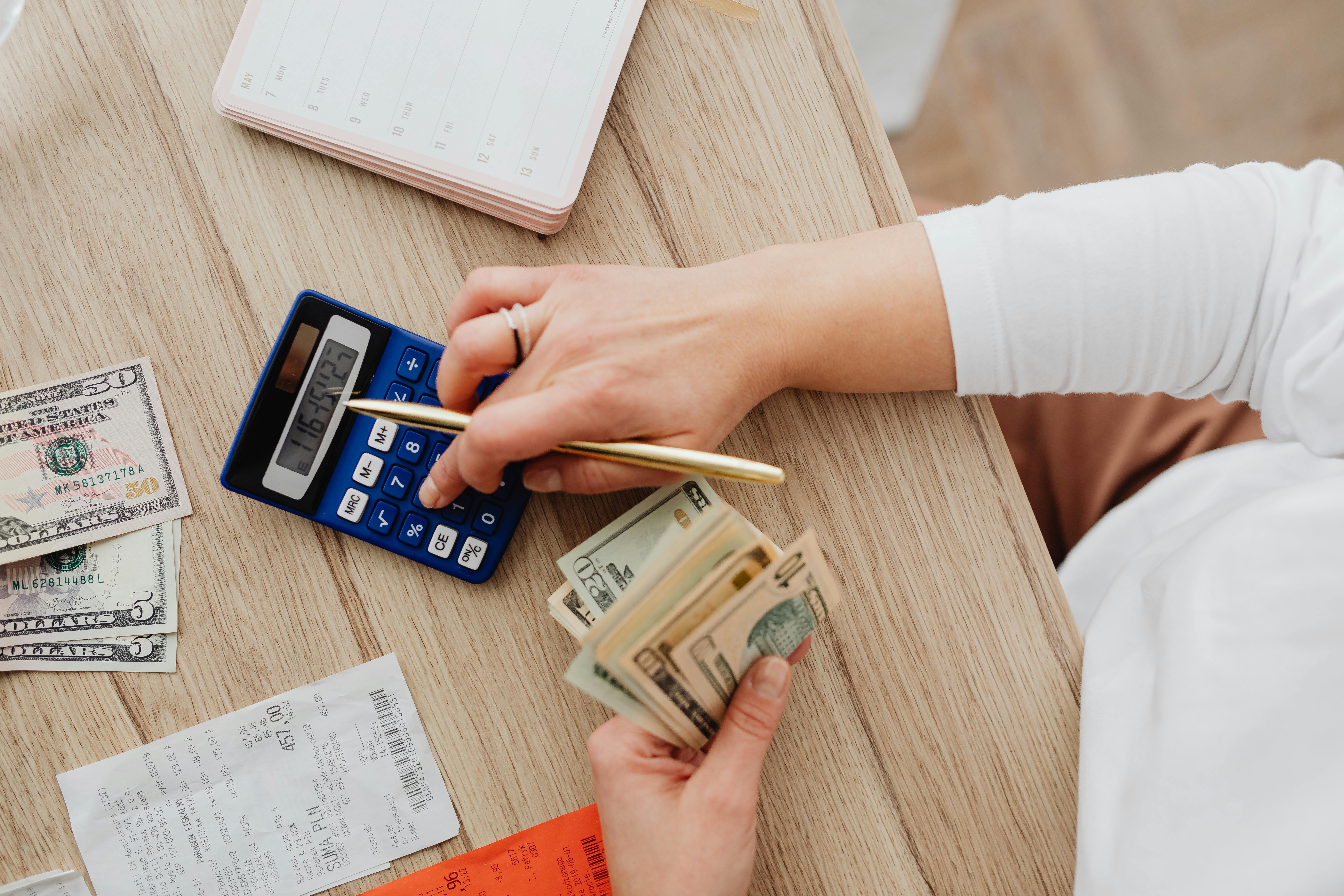 Person Using a Blue Calculator while Holding Dollar Bills