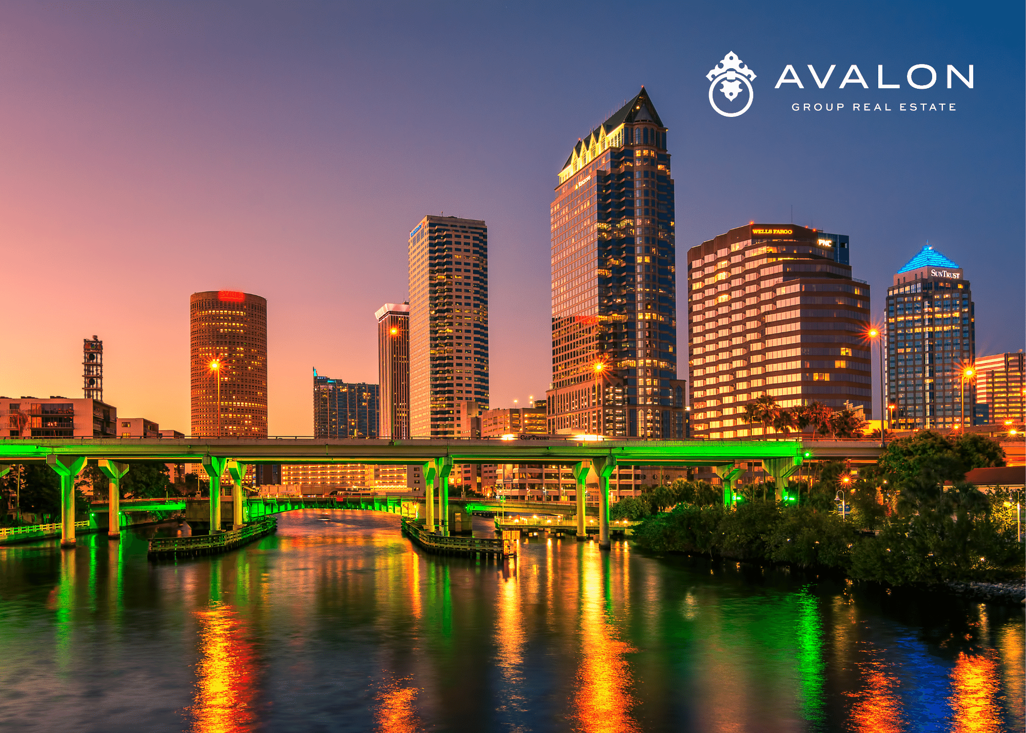 Additionally, this picture shows 6 high rise buildings in Downtown Tampa. Furthermore, the buildings have an orange tint to them because the picture is taken at sunset. There is a bridge in front of the buildings that looks green from the lighting being projected onto it.