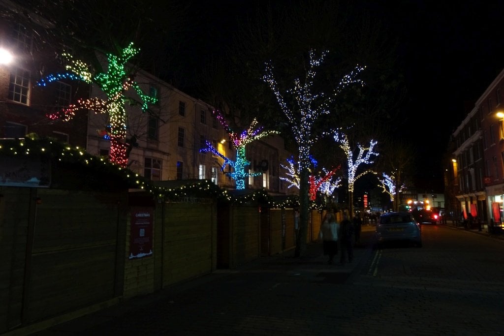 Wooden holiday market stalls trimmed with garland beside trees wrapped in colorful string lights on a downtown street at night
