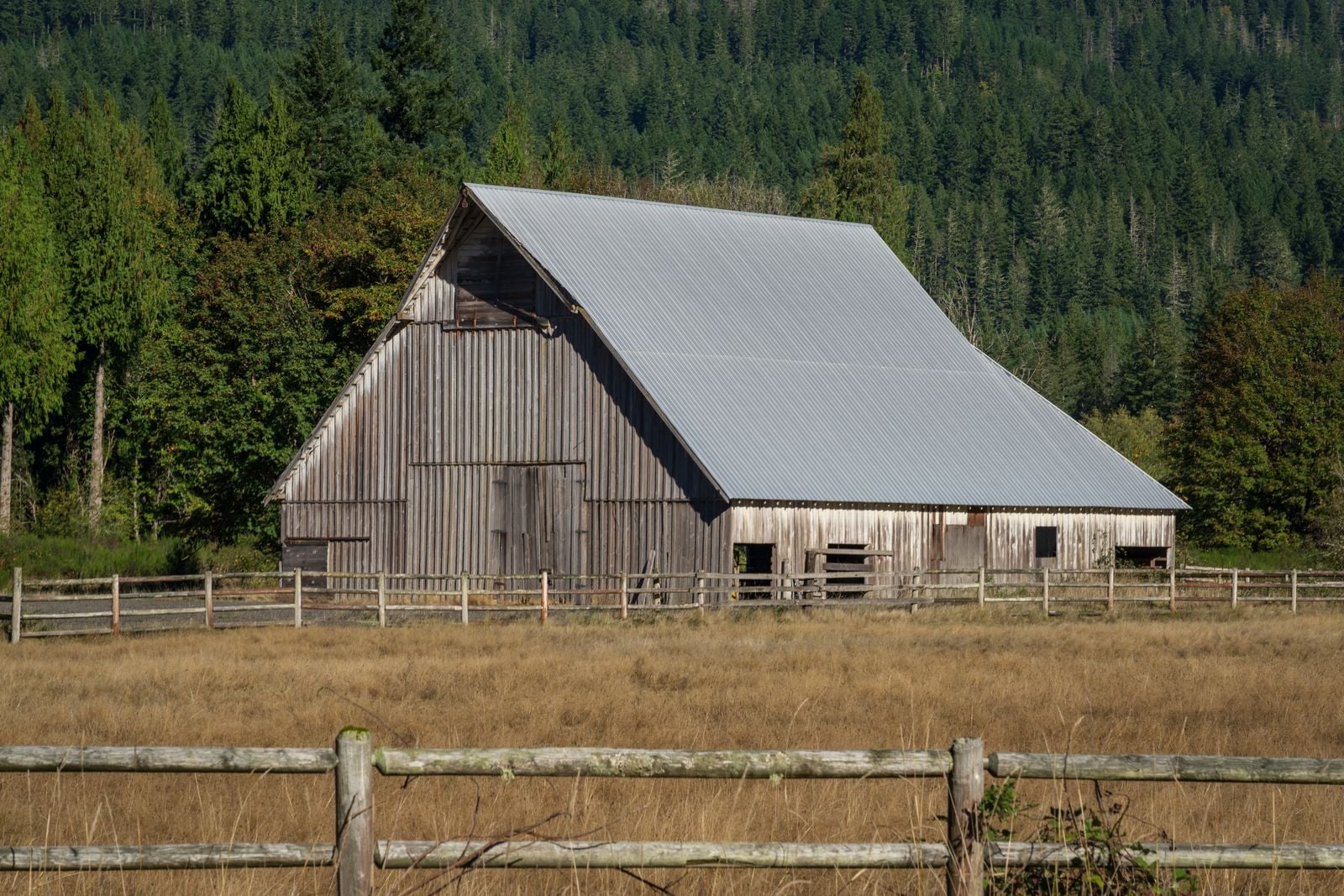 Weathered wooden barn behind a split-rail fence and dry pasture, with a forested hillside behind it