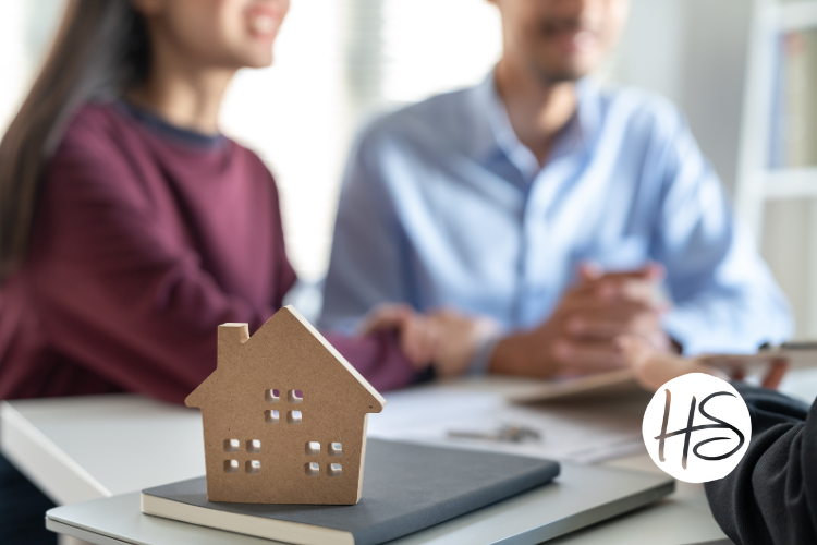 Close-up of a small wooden house model on a table in the foreground, with a couple shaking hands with a real estate agent in the background. The couple is smiling, and documents are visible on the table.