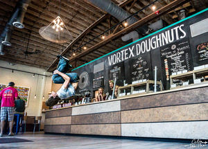 The image depicts the interior of Vortex Doughnuts, a popular doughnut shop with a lively and energetic atmosphere. The focal point is a person performing an impressive mid-air flip in front of the counter, capturing a sense of fun and excitement. The counter is lined with various doughnut options displayed under a large chalkboard menu that lists the different doughnut flavors and drinks available. The shop features an industrial design with exposed wooden beams, ductwork, and a combination of modern and rustic elements. The vibe is vibrant and welcoming, with customers and staff visible in the background, adding to the bustling ambiance.