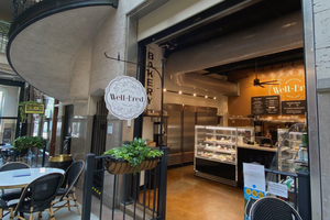 The image shows the exterior of Well-Bred Bakery & Café, located in a charming indoor setting. The bakery has an inviting storefront with a large, round hanging sign featuring the Well-Bred logo, which includes intricate decorative details and the words "Well-Bred Bakery & Café, Since 2003." Inside, you can see a display case filled with baked goods, and the interior is warm and well-lit, with a menu board on the wall. The bakery is situated next to a small outdoor seating area with a couple of tables and chairs, surrounded by plants, offering a cozy and welcoming atmosphere.