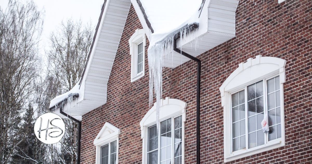 A red brick house in winter with large icicles hanging from the roof edge above white-framed windows, surrounded by a backdrop of bare trees.