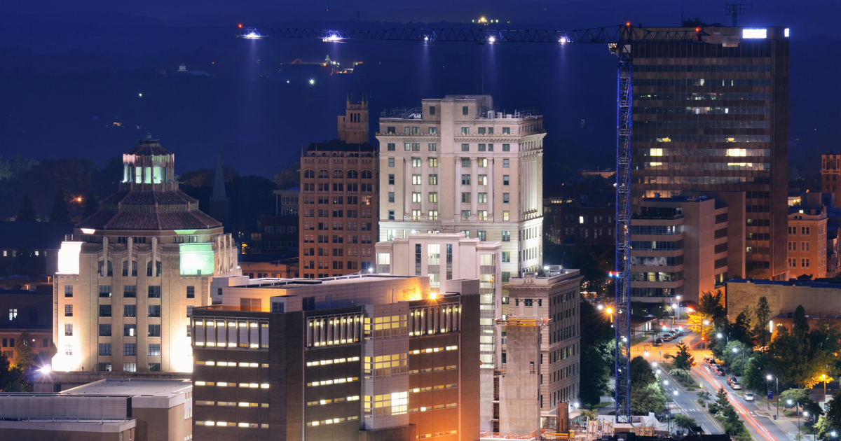 Nighttime view of downtown Asheville, North Carolina with illuminated historic buildings, modern office towers, and a construction crane against a mountain backdrop.