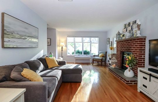 Sun-filled living room with hardwood floors, oversized picture window, and classic brick fireplace.
