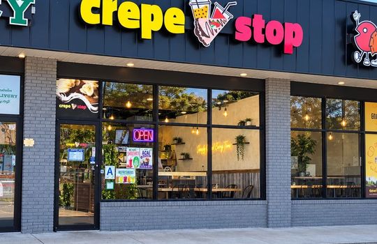 Exterior storefront of Crepe Stop in New Dorp, Staten Island, featuring colorful signage, large glass windows, outdoor parking, and the café entrance.