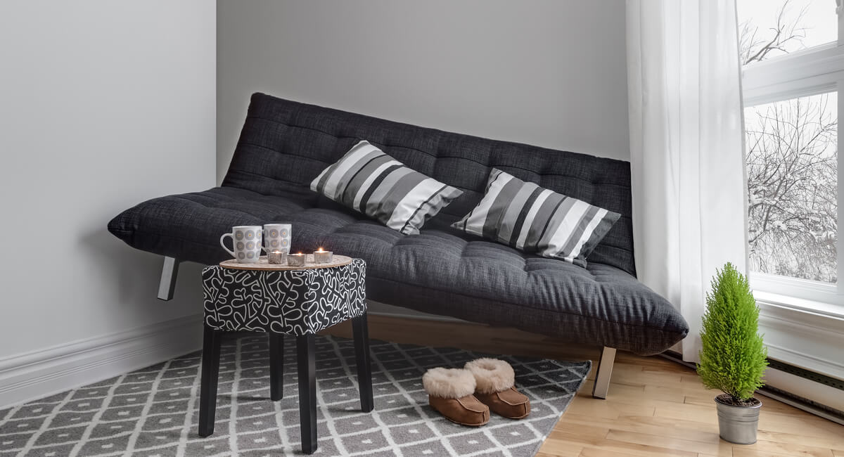 Black couch with side table and mugs and a gray rug with brown slippers on it