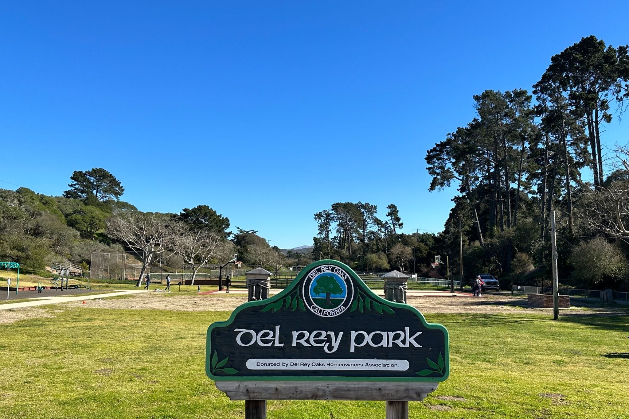 Del Rey Park sign on a sunny day, with the park lawn, oaks and Monterey pines behind it in Del Rey Oaks