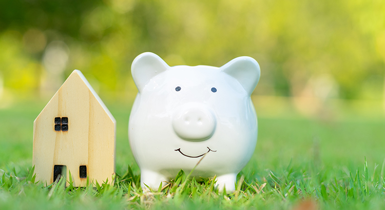 Piggy bank house figure for finance and banking or saving new home. House model and cute white piggy bank standing in green grass outdoors with copy space. Financial planning and money saving concept.