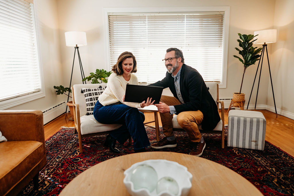 Jamie and David Quigg sitting with a client on a white couch, smiling and looking at a laptop in a warmly lit living room