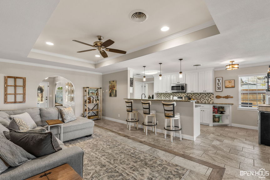 Open-concept living area with a gray sectional sofa, tiled flooring, ceiling fans, and a white modern kitchen with bar stools and stainless steel appliances.