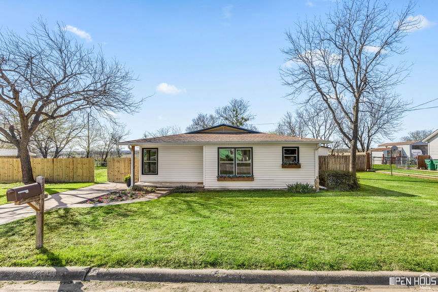 Wide-angle view of a quaint single-story home's front yard with lush green lawn and leafless tree in Jacksboro, TX.