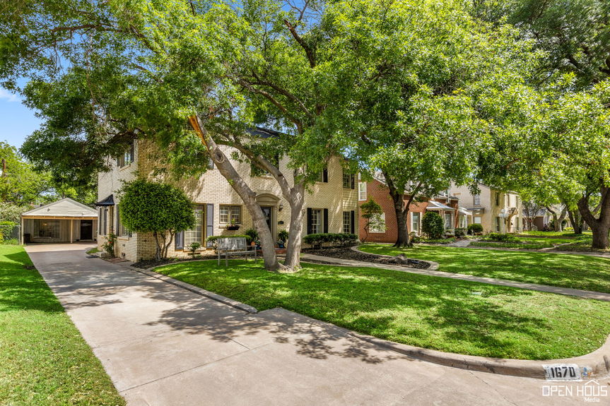 A brick home with a long driveway leading to a carport, flanked by lush trees and landscaping.