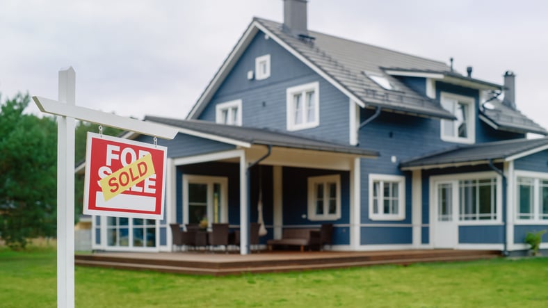 Red Property Sold Sign in a Garden in Front of a Big Blue Stylish House with Traditional Architecture. Hot Housing Market Concept with Residential Home in the Suburbs.