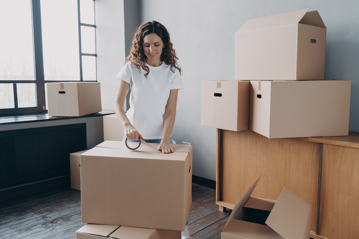 Young hispanic woman is packing containers with sticky tape. Happy mover wrapping cardboard boxes.