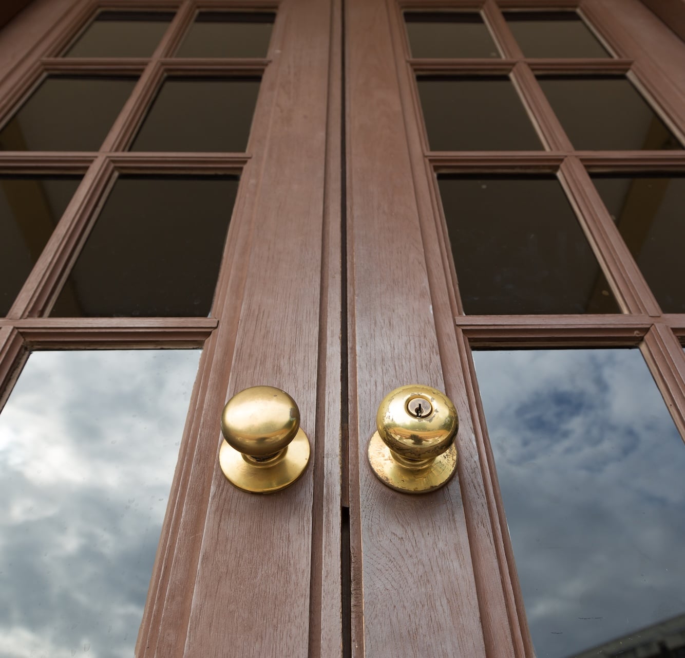 Wooden double doors with brass knobs and glass panes reflecting a cloudy sky