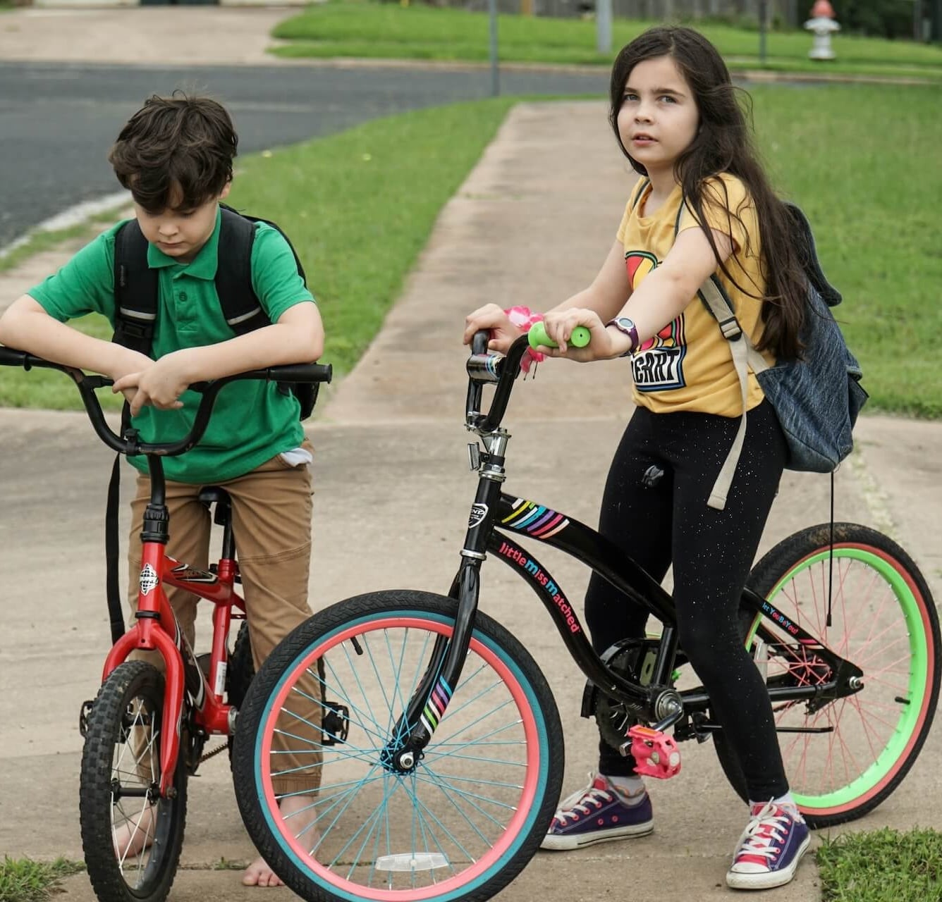 Two kids on bikes with backpacks on a suburban sidewalk in a North Pittsburgh neighborhood.