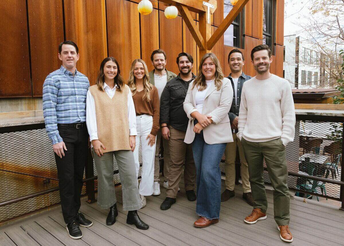 A group of eight people standing outdoors on a wooden deck with a rustic wall.