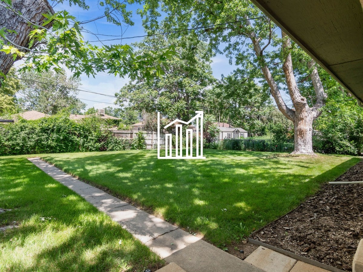 Backyard lawn with a concrete walkway, shrubs along a wood fence and tall mature shade trees under a blue sky