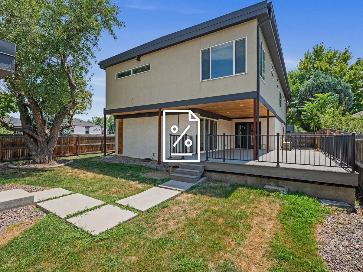 Two-story modern stucco and white brick home with a covered rear deck, black railings, a lawn with concrete stepping pads and a large shade tree