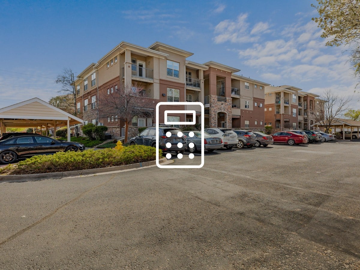 Four-story brick and stucco condominium buildings with balconies, covered parking and cars in the lot under a blue sky