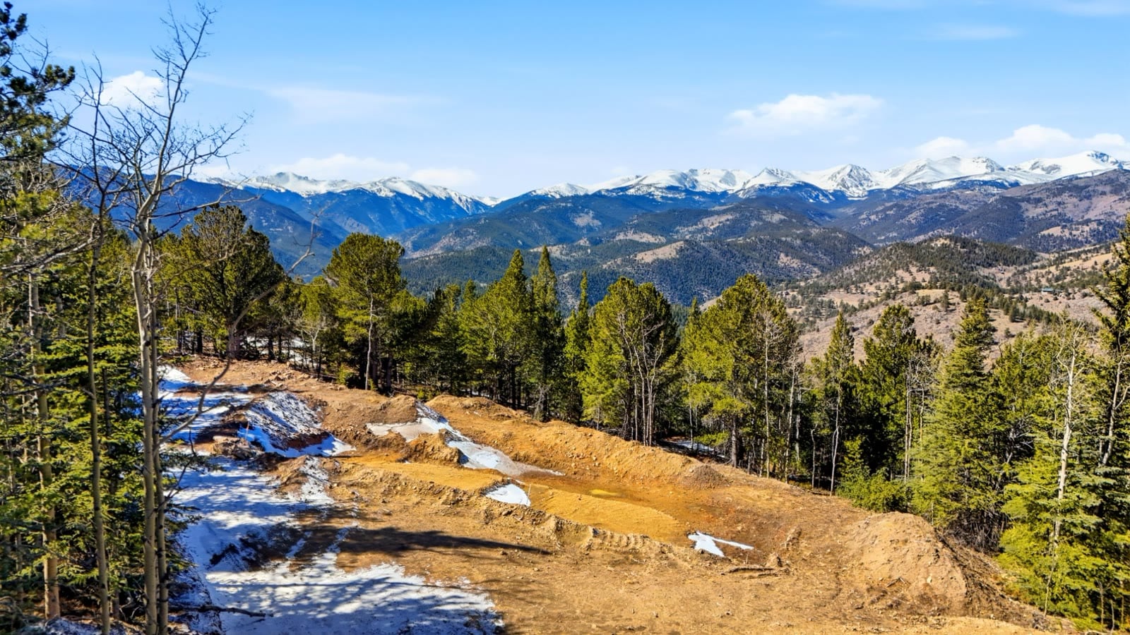 Graded building site on a mountain lot in Central City, Colorado, with patchy snow and snow-capped peaks in the distance