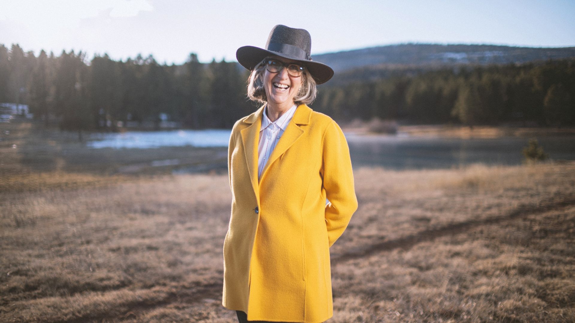 April Ralph, REALTOR in Angel Fire NM smiling and wearing a bright yellow coat and a wide-brimmed hat, stands in in front of Monte Verde lake.