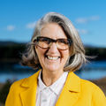 April Ralph, wearing a bright yellow coat and glasses, smiling with a scenic mountain lake in the background.