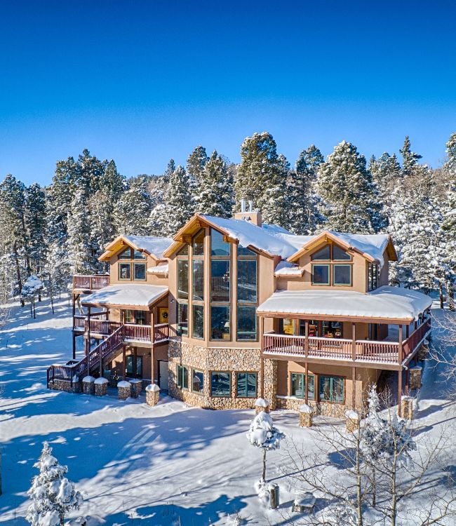 A spacious mountain home, featuring soaring glass windows, surrounded by snow and pine trees.