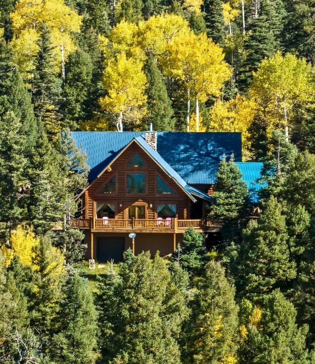 A stunning log cabin home with a blue roof, surrounded by lush green pine trees and golden aspens in Angel Fire, New Mexico.