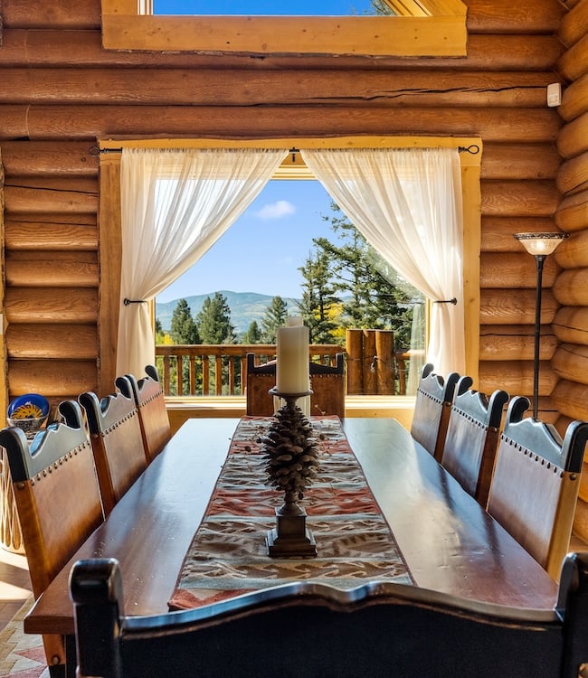 A rustic log cabin dining room with a large wooden table, decorative pinecone centerpiece, and curtains framing a breathtaking mountain view.