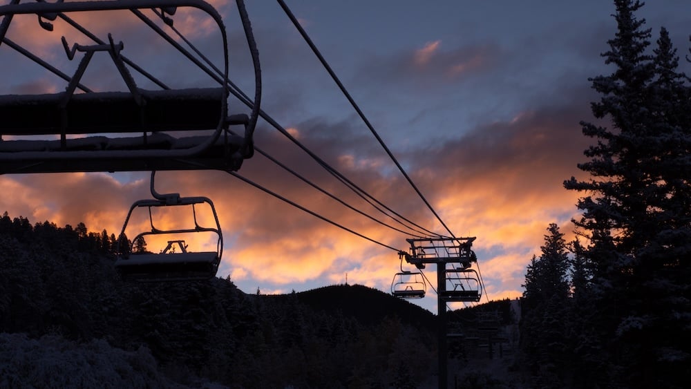 Silhouette of ski lift chairs at Angel Fire Resort against a vivid sunset sky, surrounded by mountain trees.