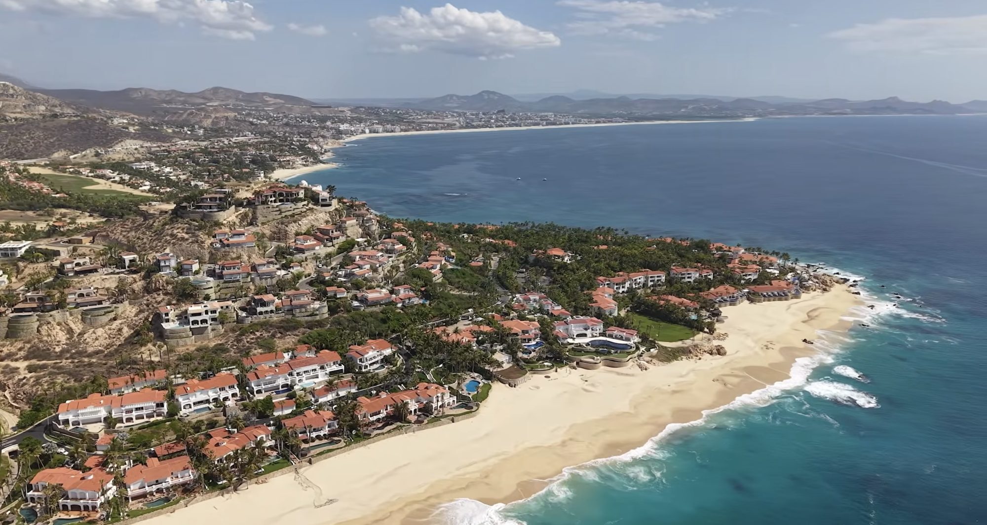 Aerial view of Palmilla: red-tiled residences stepping down the headland to a crescent of sand, with the Sea of Cortez and San Jos&eacute; del Cabo beyond.